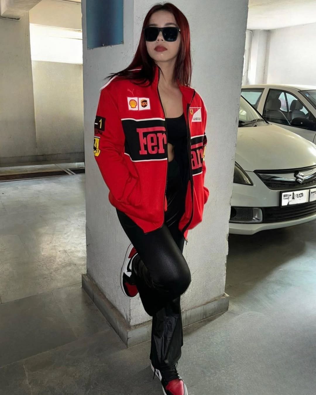 Woman posing in parking area wearing red Ferrari racing jacket, black leather pants, and sneakers, representing premium F1 apparel by ALTR.